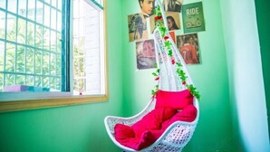 Une pièce avec un mur vert, un fauteuil suspendu blanc agrémenté de coussins rouges, et une fenêtre donnant sur un paysage verdoyant.