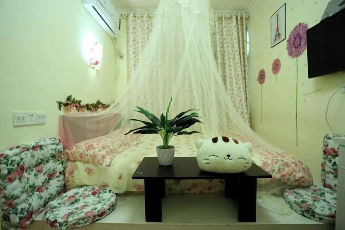 Una habitación con una cama, una mesita con una planta, un cojín con forma de gato y una mosquitera.