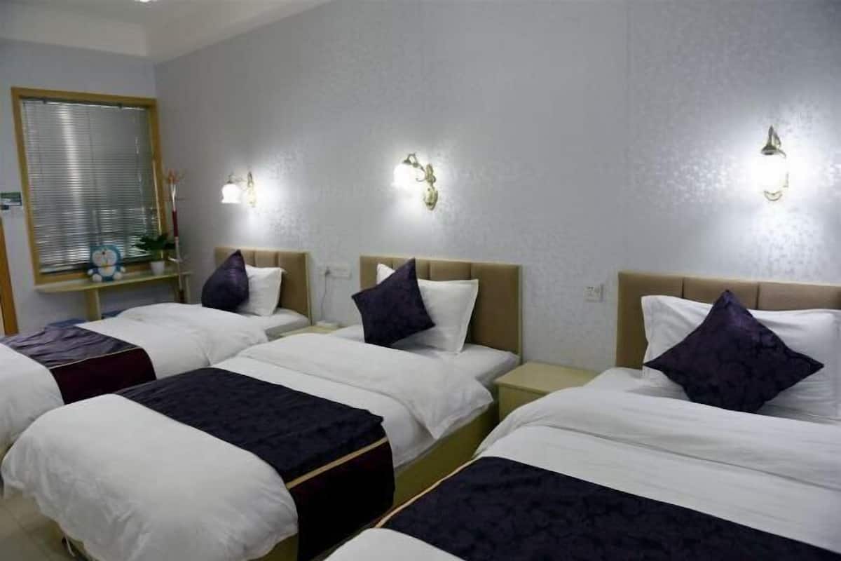 Habitación de hotel con tres camas, cada una con ropa blanca y cabeceras oscuras. Hay luces fijadas a la pared y una ventana con persianas.