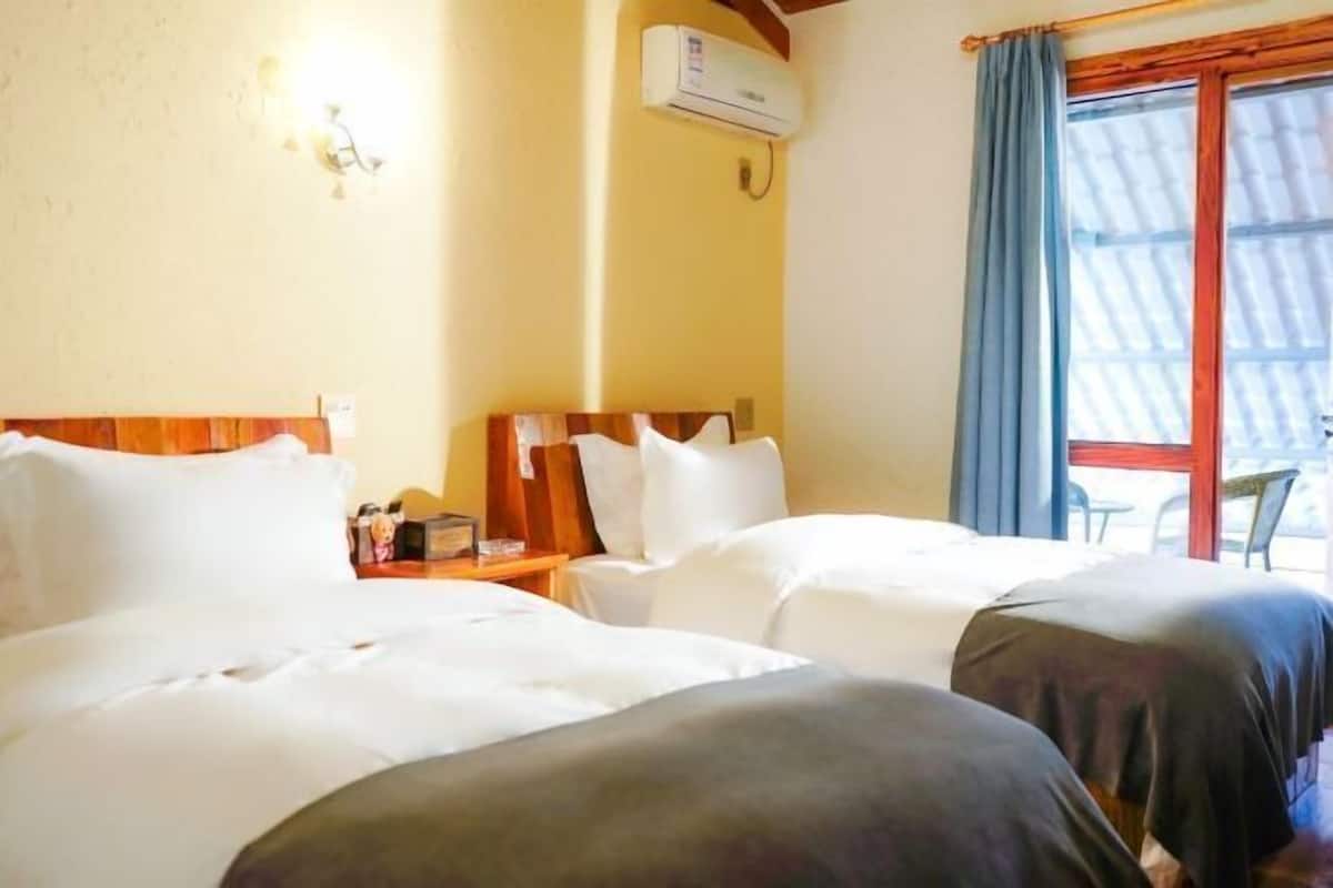 Habitación de hotel con dos camas, cabecera de madera, equipo de aire acondicionado, una mesita con lámpara y ventana con cortinas.