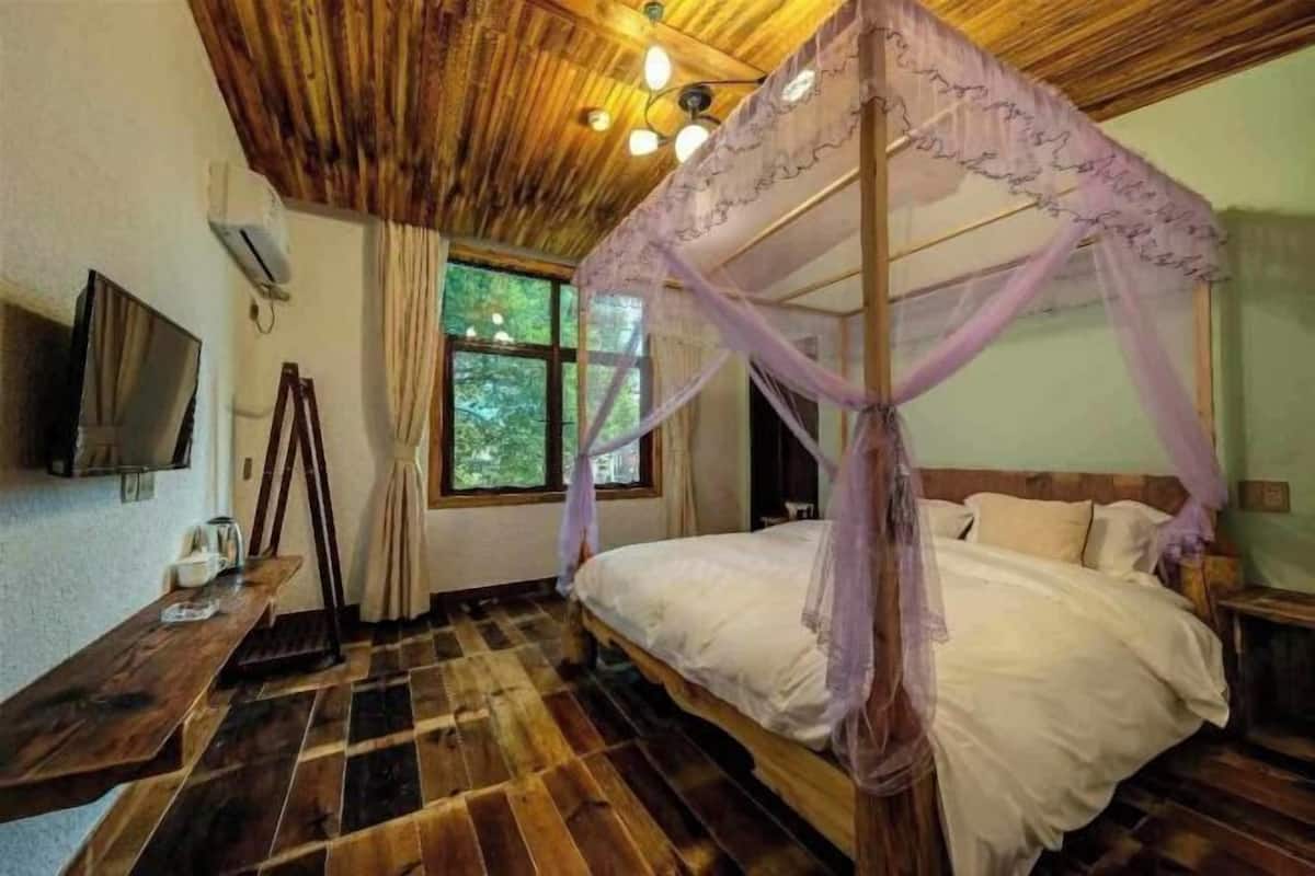 Un dormitorio con techo de madera, cama con dosel y mosquitera, televisor en la pared y ventana con vista a árboles.