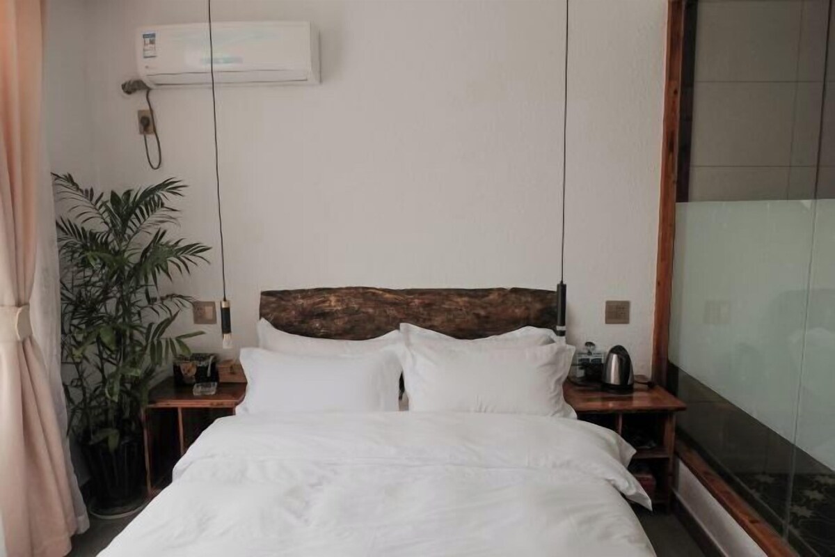 A hotel room with a bed, a nightstand, a potted plant, a wooden headboard, and an air conditioning unit.