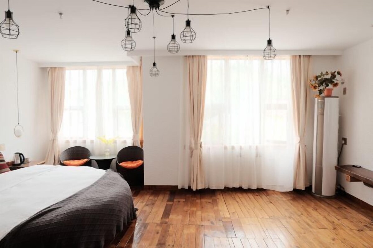 A hotel room with a bed, two chairs, a desk, a lamp, a plant, and a wardrobe.