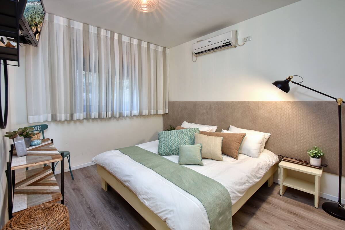 Quarto com cama, mesas de cabeceira, luminária, janela com cortinas e ar-condicionado.