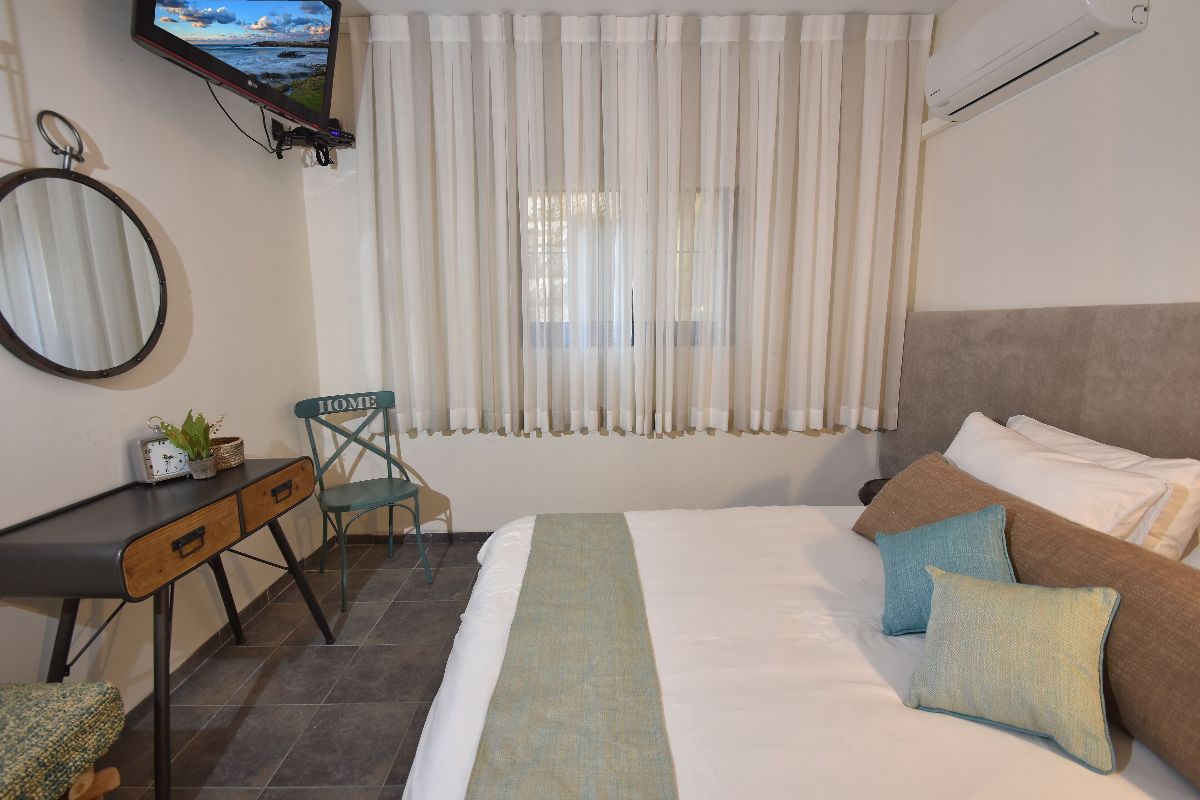 Un dormitorio con cama, almohadas, un escritorio con silla, televisor y ventana con cortinas.