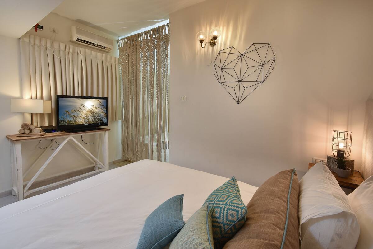 Quarto com cama, televisão em suporte, luminária fixada na parede e um quadro de parede com padrão geométrico.