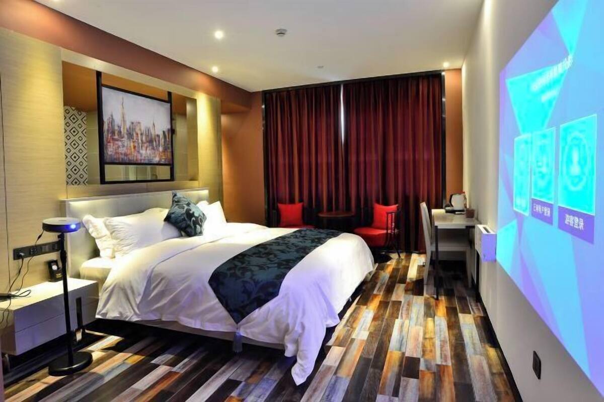 Une chambre d’hôtel moderne, dotée d’un grand lit, d’un bureau et d’une chaise. Un téléviseur est fixé au mur, et un écran de projection affiche des informations.