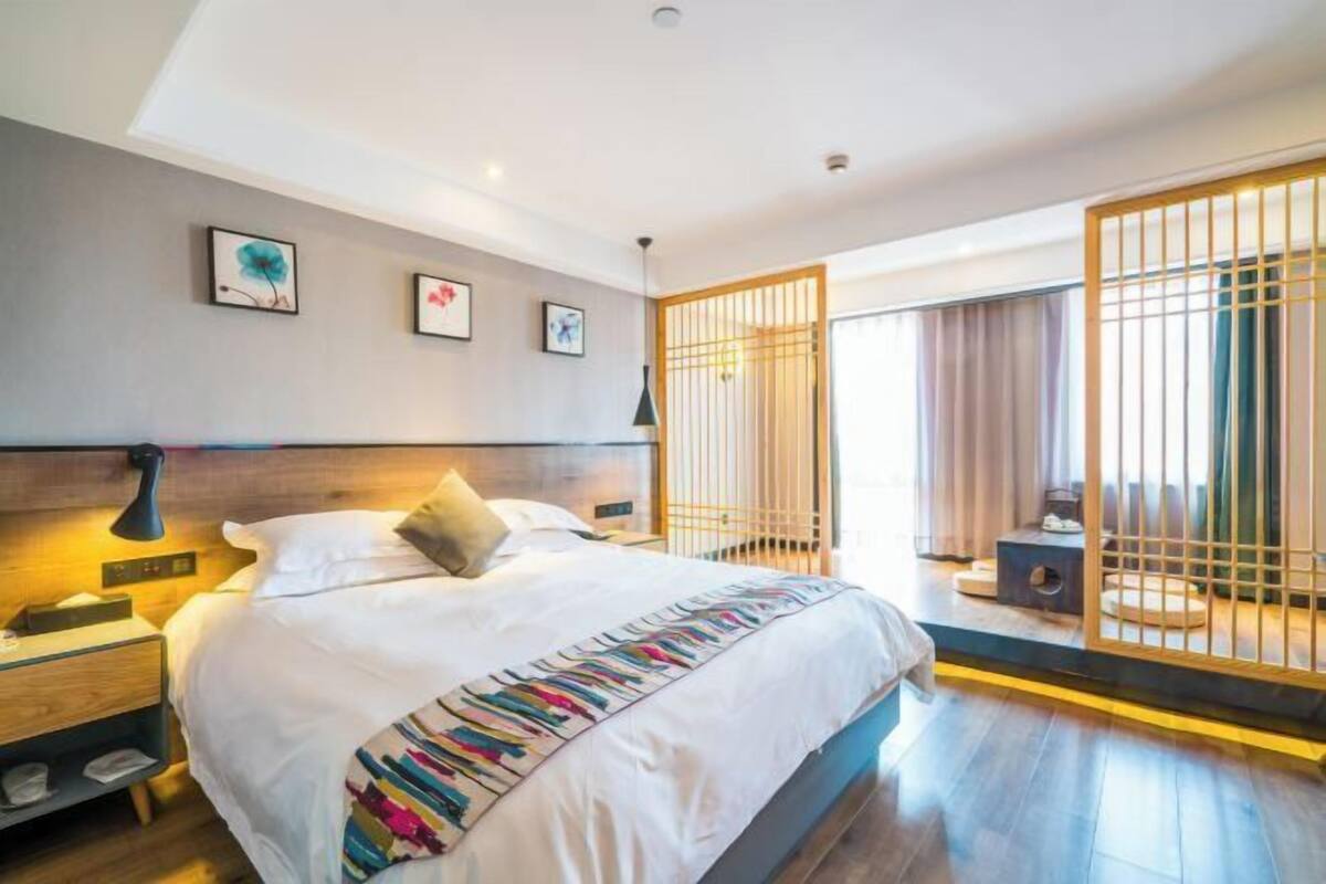 Une chambre d’hôtel moderne dotée d’un grand lit, de tables de chevet, d’une petite table, d’une fenêtre avec des rideaux et d’un paravent décoratif.