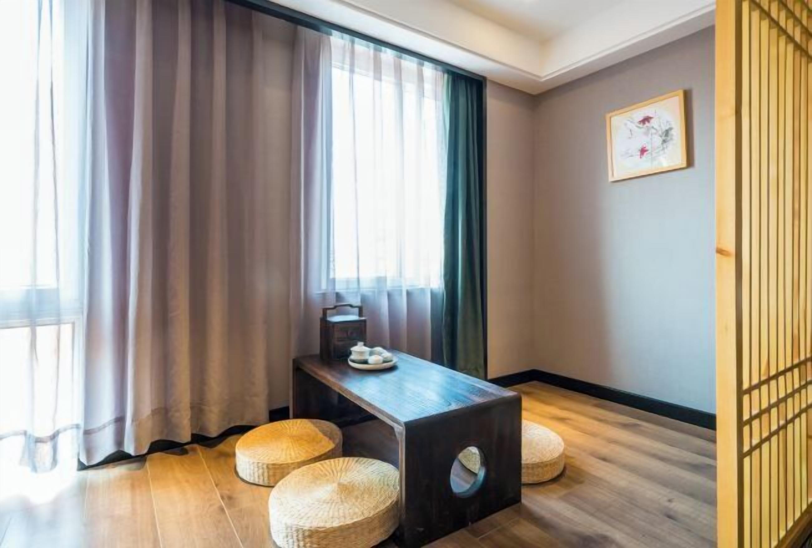 Una habitación con una mesa de madera, dos pufs tejidos, una ventana con cortinas translúcidas y un cuadro en la pared.