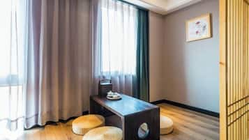 Una habitación con una mesa de madera, dos pufs tejidos, una ventana con cortinas translúcidas y un cuadro en la pared.