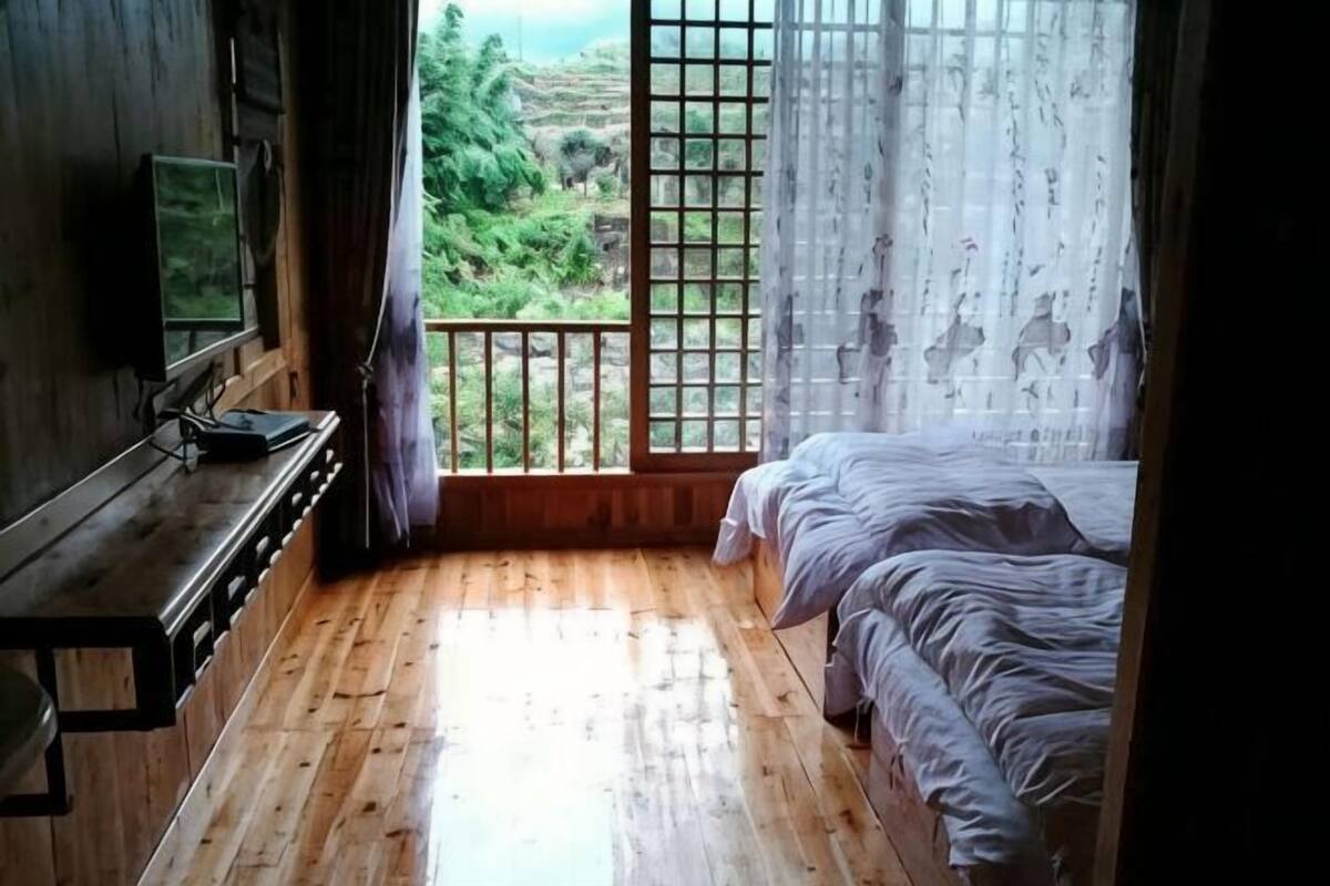Quarto com piso de madeira, cama com roupa de cama branca, TV sobre um suporte de madeira e vista para o verde através de uma janela com grade de metal.