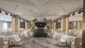 Un salón de banquetes con mesas redondas, sillas blancas y un arco floral en el centro.