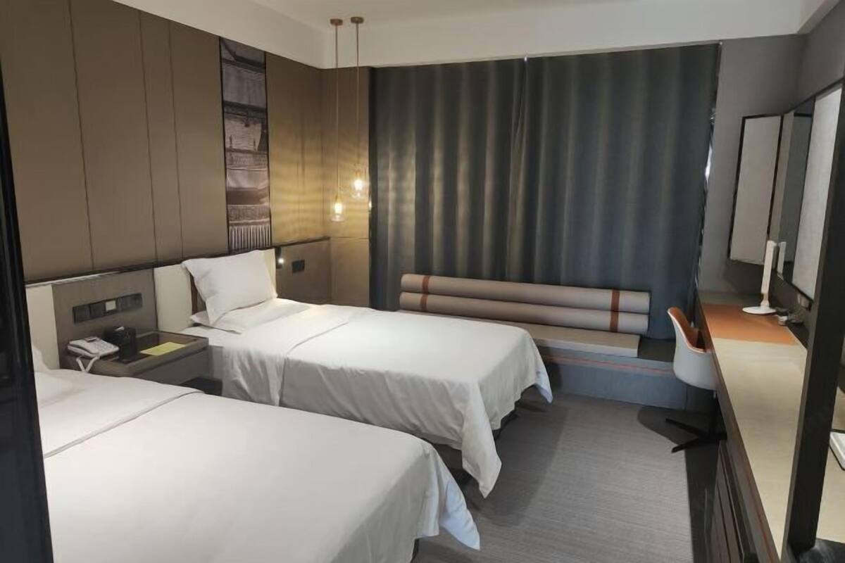 Habitación de hotel con dos camas individuales, un escritorio con silla, un espejo grande y una zona de estar con un banco.