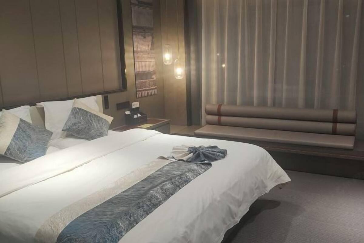 Una cama bien tendida con una colcha estampada, una mesita de noche con una lámpara y una ventana con cortinas.