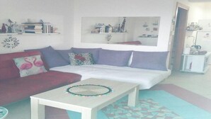 Sala de estar com sofá, mesa de centro e uma pequena mesa com objetos decorativos. Há um espelho na parede e uma geladeira ao fundo.