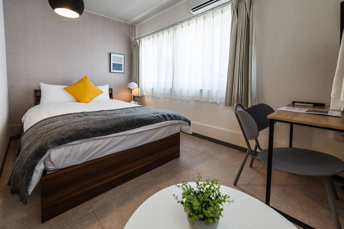 Una camera d'albergo moderna con un letto, una scrivania, una sedia, una lampada e una pianta in vaso.