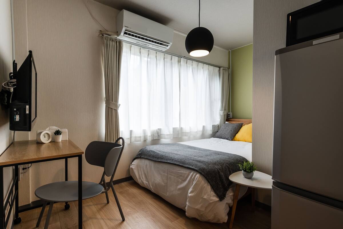 Una camera d'albergo compatta con un letto, una scrivania e una sedia, un piccolo tavolino con una pianta, un condizionatore d'aria e una televisione.