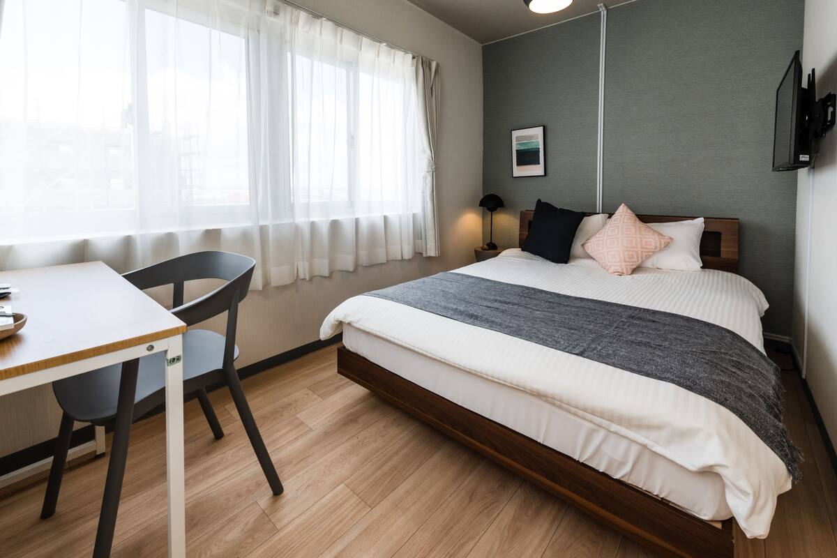 Una camera d'albergo con un letto, una scrivania, una sedia, una finestra con tende, una TV a parete e una lampada da comodino.
