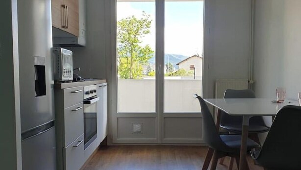 A modern kitchen with a refrigerator, microwave, and oven. There is a dining table with chairs and a view of the outdoors through a large window. - 3 rooms Grenoble - Saint Martin d'heres (Saint-Martin-d'Hères)