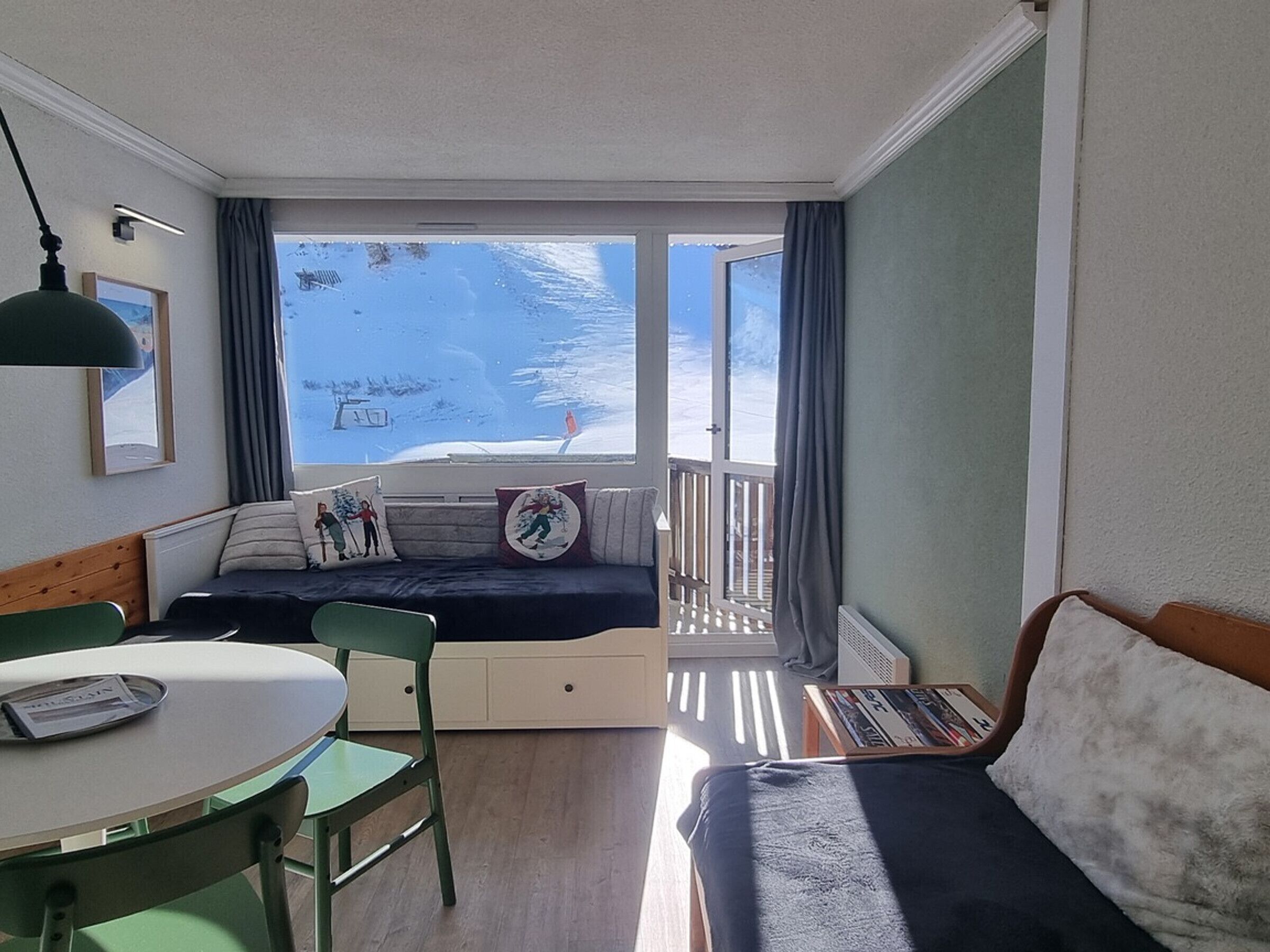 Una habitación con balcón, sofá, mesa de comedor con sillas y ventana con vista a un paisaje nevado.