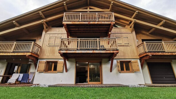 A two-story wooden building with balconies, a covered entrance, and a grassy front yard. - Cozy T3 apartment near center with terrace and parking (Samoëns)