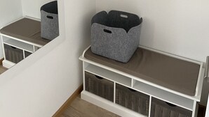 Una habitación con pared blanca, un espejo, un banco blanco con compartimentos de almacenamiento y un cesto de almacenamiento gris.