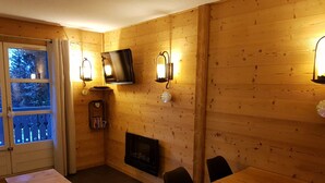 Ein gemütlicher Raum mit Holzwänden, einem Kamin, einem darüber montierten Fernseher und einem Essbereich mit Tisch und Stühlen.