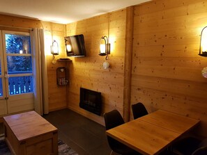 Ein gemütlicher Raum mit Holzwänden, einem Kamin, einem darüber montierten Fernseher und einem Essbereich mit Tisch und Stühlen.