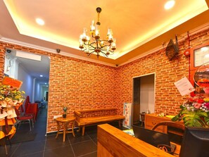 A hotel lobby with a brick wall, a chandelier, a reception desk, and a dining area with chairs and a bench.