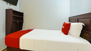 Una cama individual con una colcha roja y blanca, dos almohadas, un cabecero de madera y un televisor montado en la pared.