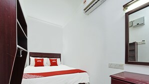 Una cama bien hecha con una colcha blanca y roja, un cabecero de madera y dos almohadas rojas con el texto "OYO". Hay una mesita de noche de madera con un cajón, un espejo en la pared y una unidad de aire acondicionado sobre la cama.