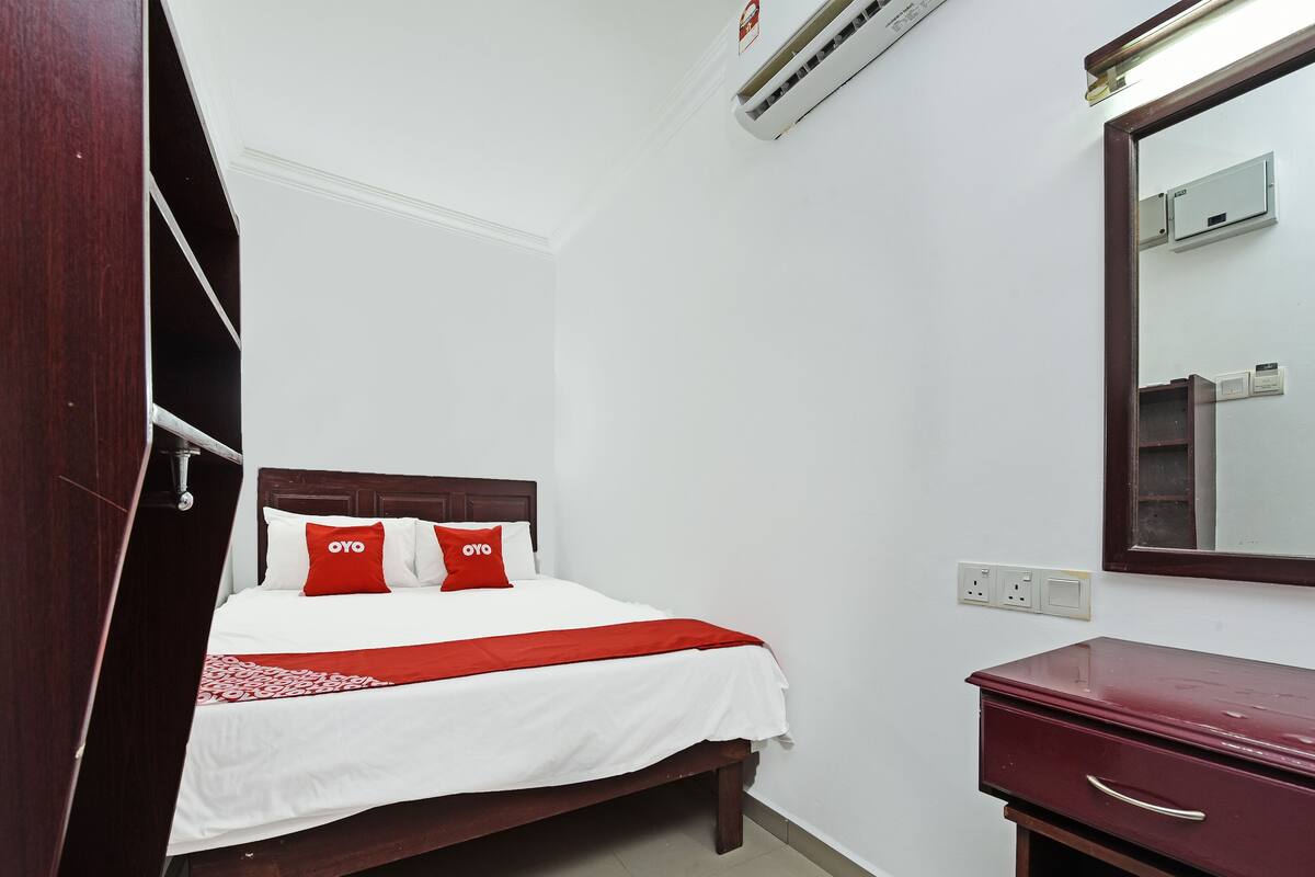 Una cama bien hecha con una colcha blanca y roja, un cabecero de madera y dos almohadas rojas con el texto "OYO". Hay una mesita de noche de madera con un cajón, un espejo en la pared y una unidad de aire acondicionado sobre la cama.