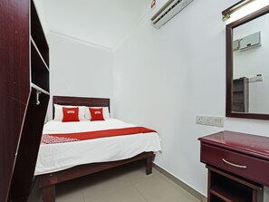 Una cama bien hecha con una colcha blanca y roja, un cabecero de madera y dos almohadas rojas con el texto "OYO". Hay una mesita de noche de madera con un cajón, un espejo en la pared y una unidad de aire acondicionado sobre la cama.
