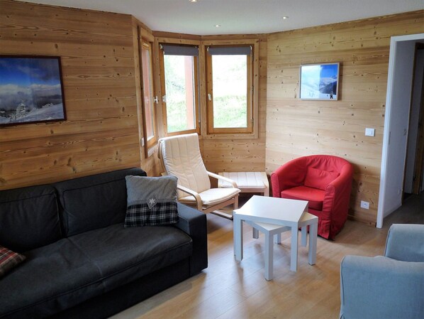 A cozy room with a wooden interior, a black sofa, a red chair, a white table, and two windows providing natural light.