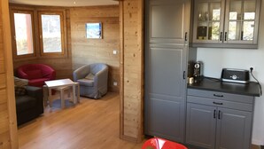 Interior acolhedor de uma cabana com piso de madeira, área de refeições com mesa e cadeiras, cozinha com armários e eletrodomésticos, e uma janela com vista.