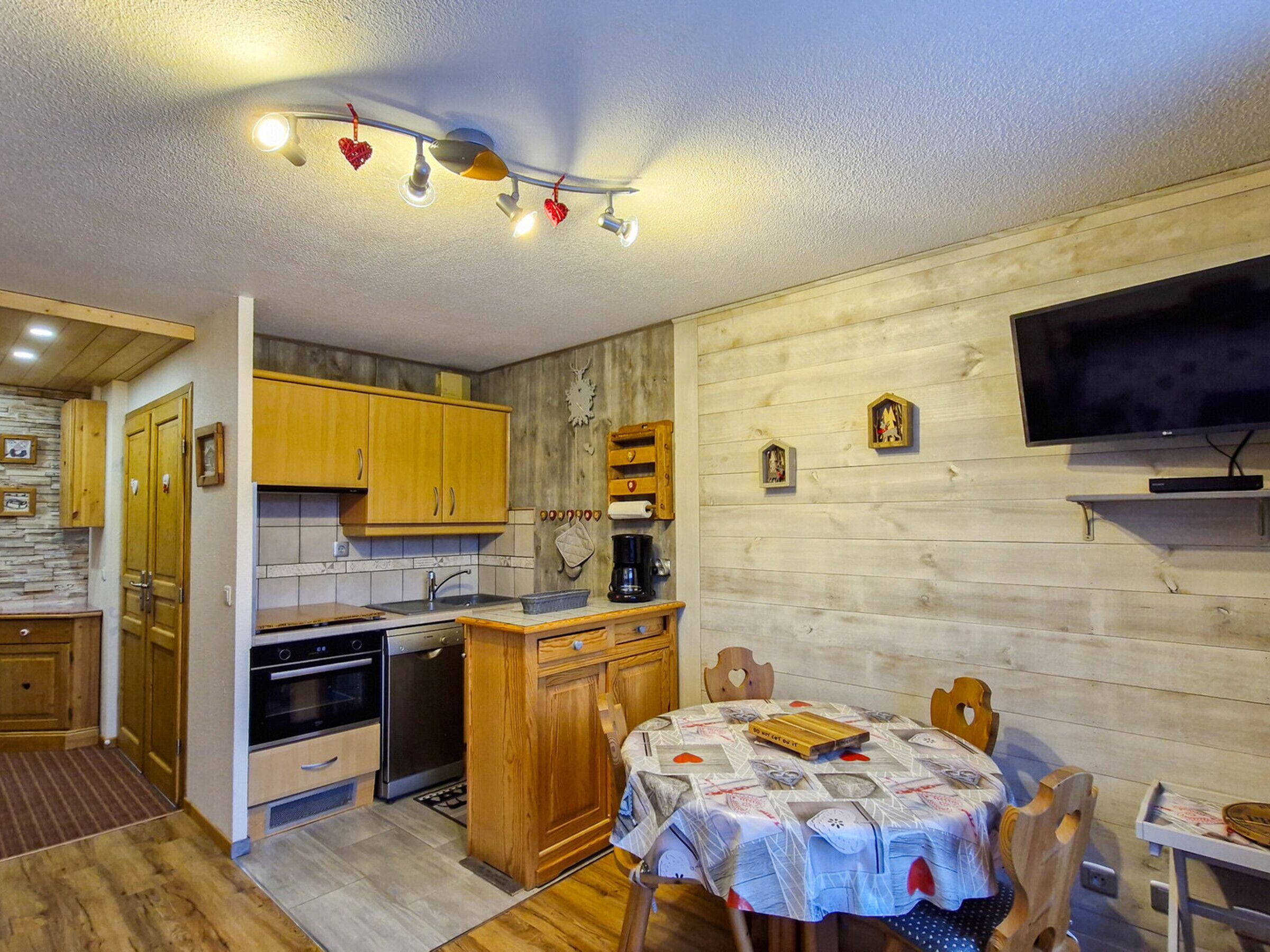 A compact kitchen with wooden cabinets, a dining table with a patterned tablecloth, and a TV mounted on the wall.