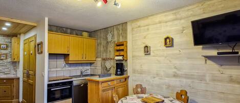A compact kitchen with wooden cabinets, a dining table with a patterned tablecloth, and a TV mounted on the wall.