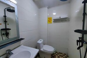 Un baño pequeño con lavamanos, inodoro y ducha. Hay un cartel en la pared y un perchero encima del inodoro.