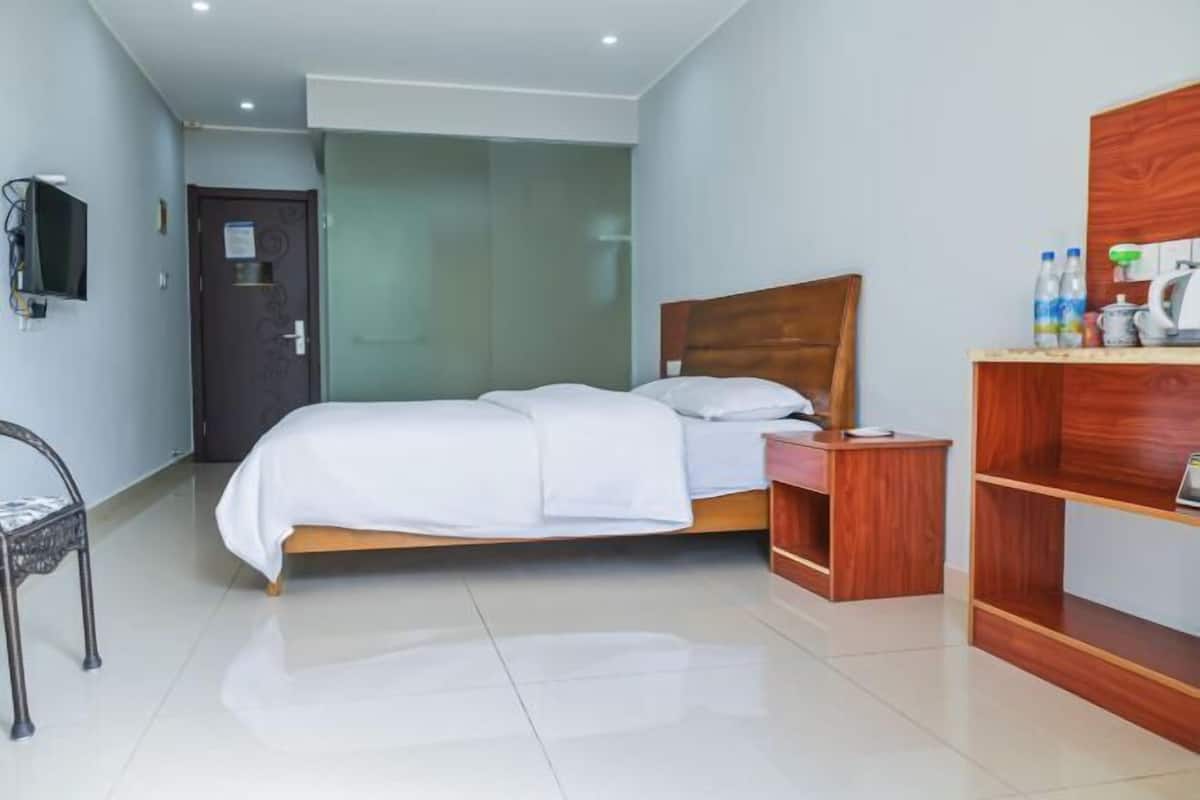 Une chambre d’hôtel avec un lit, une table de chevet en bois, une télévision et une étagère avec des bouteilles d’eau.