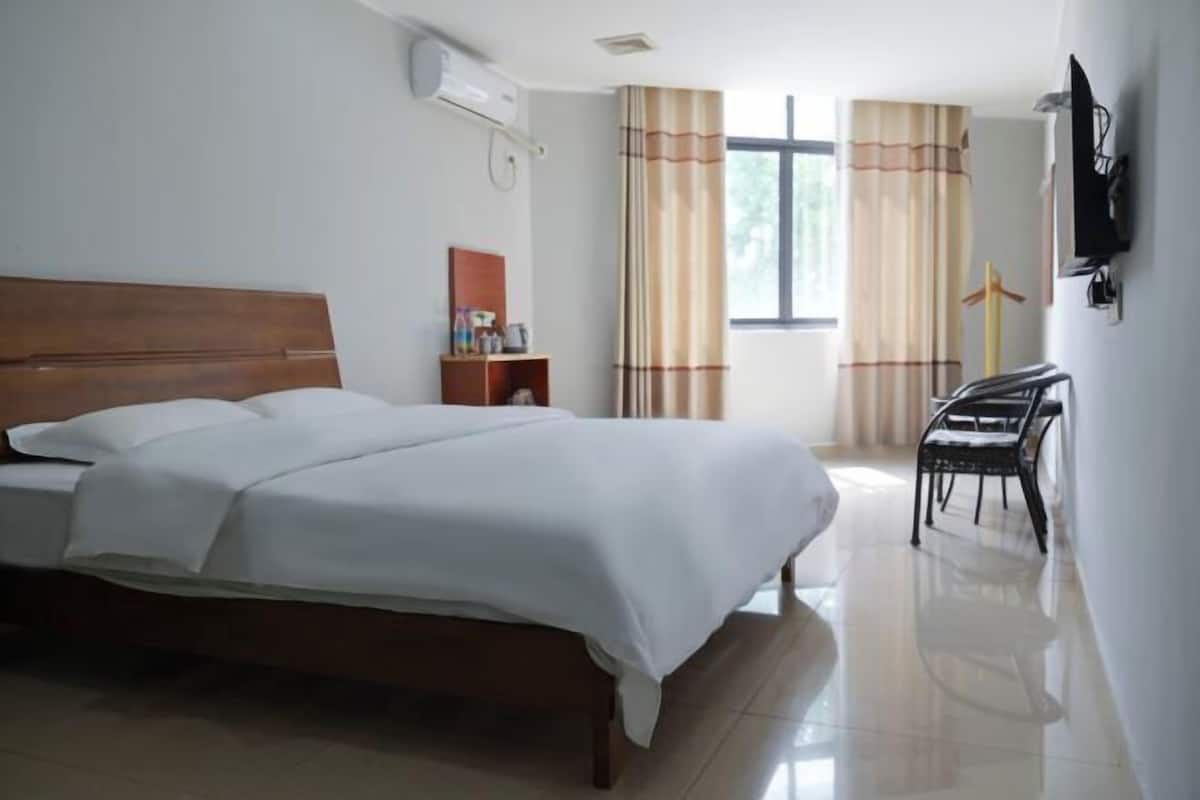 Une chambre d’hôtel avec un lit, une petite table de chevet, une chaise, une télévision et une fenêtre avec des rideaux.