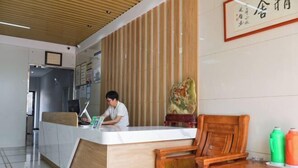 Un mostrador de recepción con una persona sentada, una silla de madera y una mesa con dos botellas.