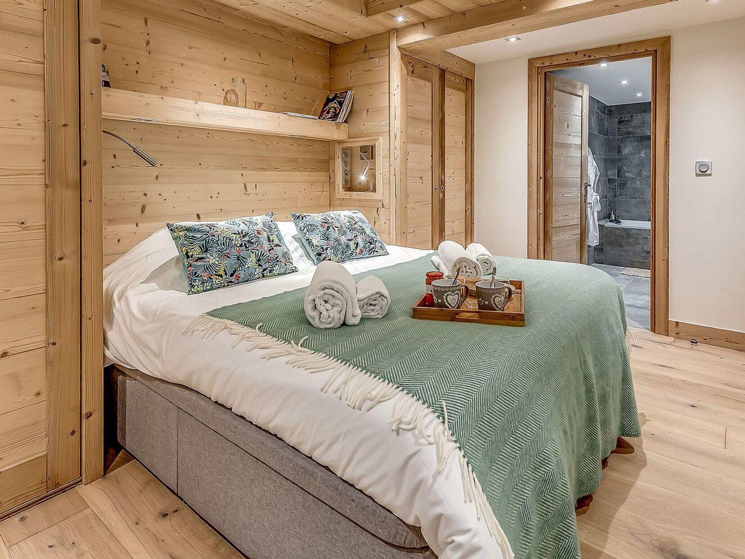 A wooden room with a bed, pillows, a tray with towels and mugs, and a view of a stone wall.