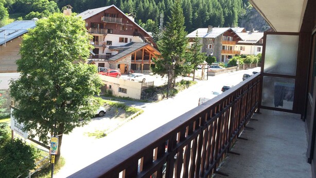 Un chalet de montagne composé de plusieurs bâtiments, doté d’un balcon avec une balustrade en bois et offrant une vue sur une route où sont garées des voitures.
