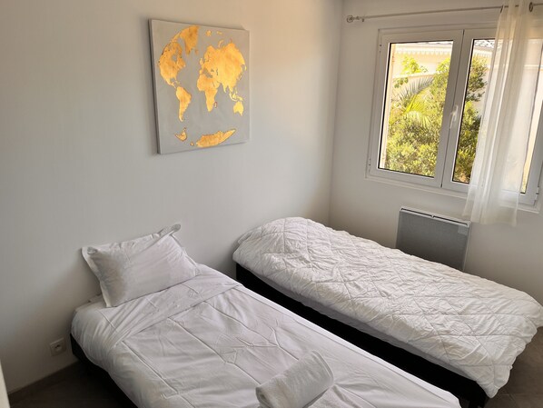 Une chambre avec deux lits simples, une œuvre d’art représentant une carte du monde accrochée au mur et une fenêtre donnant sur un paysage verdoyant.