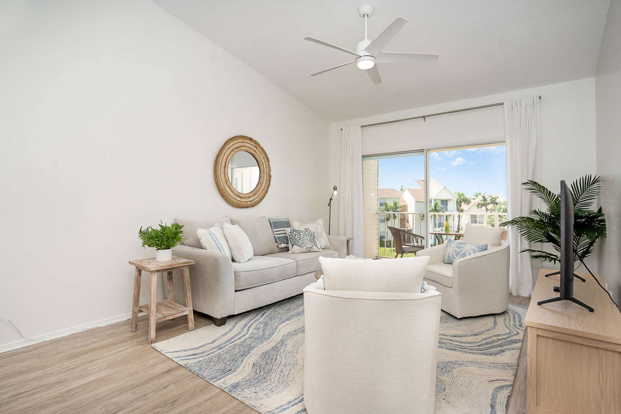 Una sala de estar moderna con un sofá, un sillón y una mesa de centro, un ventilador de techo y un gran ventanal con vistas a un barrio residencial.
