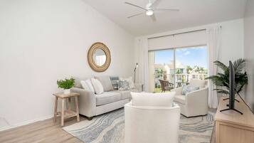 Una sala de estar moderna con un sofá, un sillón y una mesa de centro, un ventilador de techo y un gran ventanal con vistas a un barrio residencial.