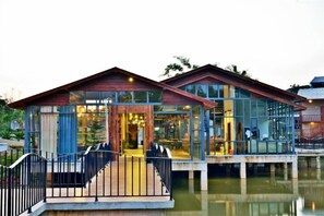 Une propriété située au bord de l’eau, dotée d’un bâtiment entièrement vitré, d’une terrasse en bois et d’une rambarde en métal.