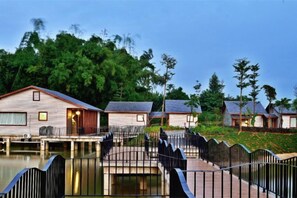 A row of wooden cabins with a bridge connecting them over a body of water, surrounded by greenery.