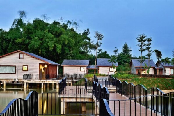 A row of wooden cabins with a bridge connecting them over a body of water, surrounded by greenery.