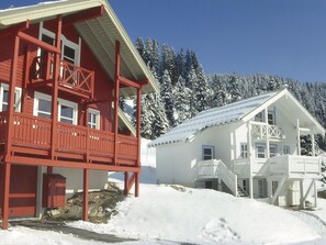 A red and white cabin with a balcony and a covered porch, surrounded by snow-covered trees.